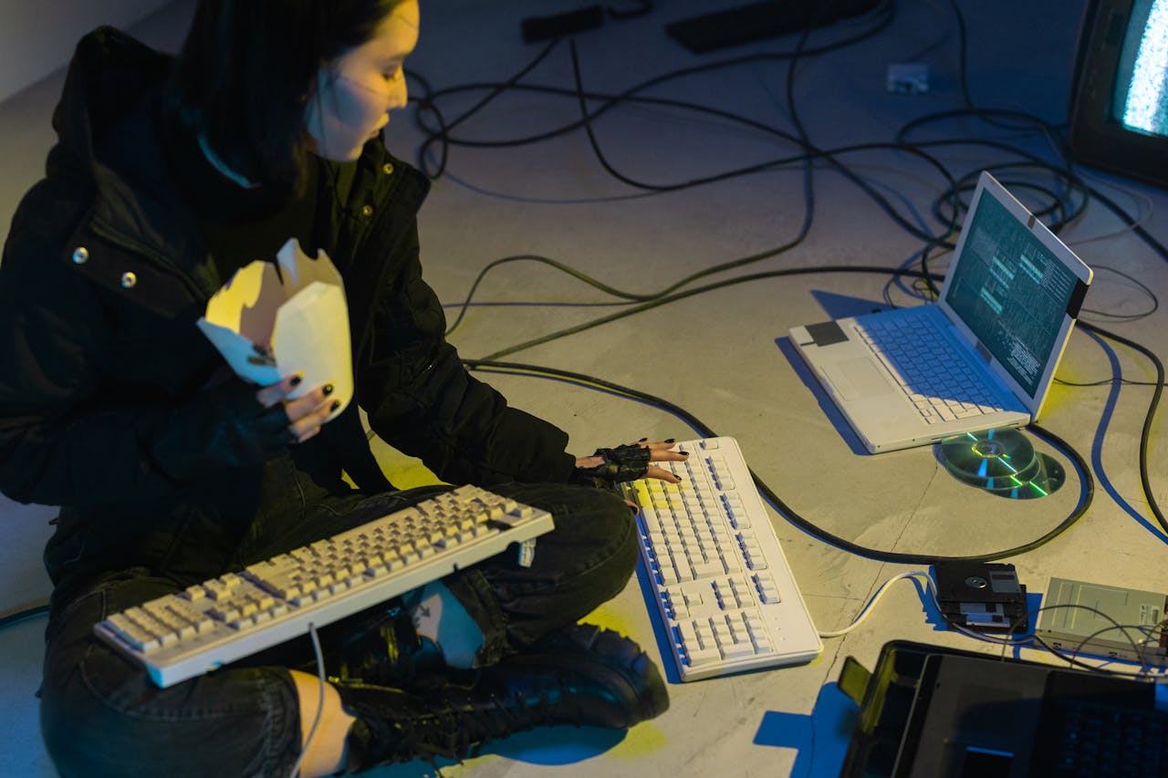 A young woman in a dark room surrounded by computers and cables, eating and typing on keyboards.